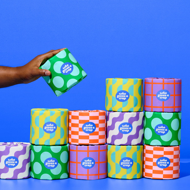 Hand holding a Who Gives A Crap toilet paper roll in front of stacked colourful rolls on bright blue background