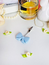 Light blue silicone bow J clip displayed on a white surface alongside pearls, perfume, and small fabric flowers, showing the clip’s delicate, feminine design and metal claw tip.