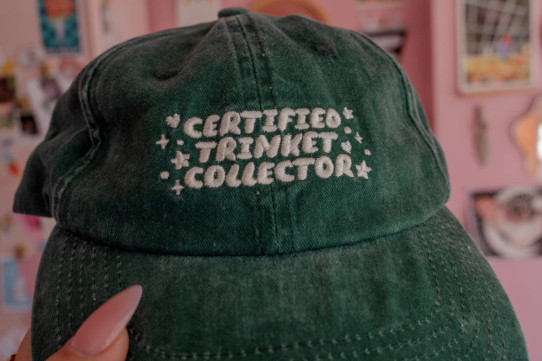 Hunter green baseball cap with embroidered text reading “Certified Trinket Collector,” front view. Hunter green baseball cap with embroidered text reading “Certified Trinket Collector,” front view.