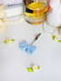 Light blue silicone bow J clip displayed on a white surface alongside pearls, perfume, and small fabric flowers, showing the clip’s delicate, feminine design and metal claw tip.