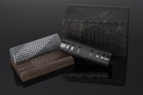 Two polished cylinders—one black shungite, one dark grey soapstone—standing upright on a solid oak stand engraved with the Flower of Life pattern.