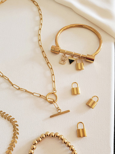Collection of Elegant Gold Jewelry Laying in Display. 1 Gold Round Paperclip Toggle Necklace Chain.  1 Gold Arrow Custom Necklace Chain. 1 Gold Bead Ball Bracelet. 3 Small Gold Plated Stainless Steel Padlock Charms. 1 Gold Stainless Steel Hope Bracelet Bar with 3 Bracelet Charms; 1 Cubic Zirconia Initial Letter Charm, 1 Dainty Black and Gold Marble Arrow Charm, 1 Gold Stainless Steel Padlock Charm