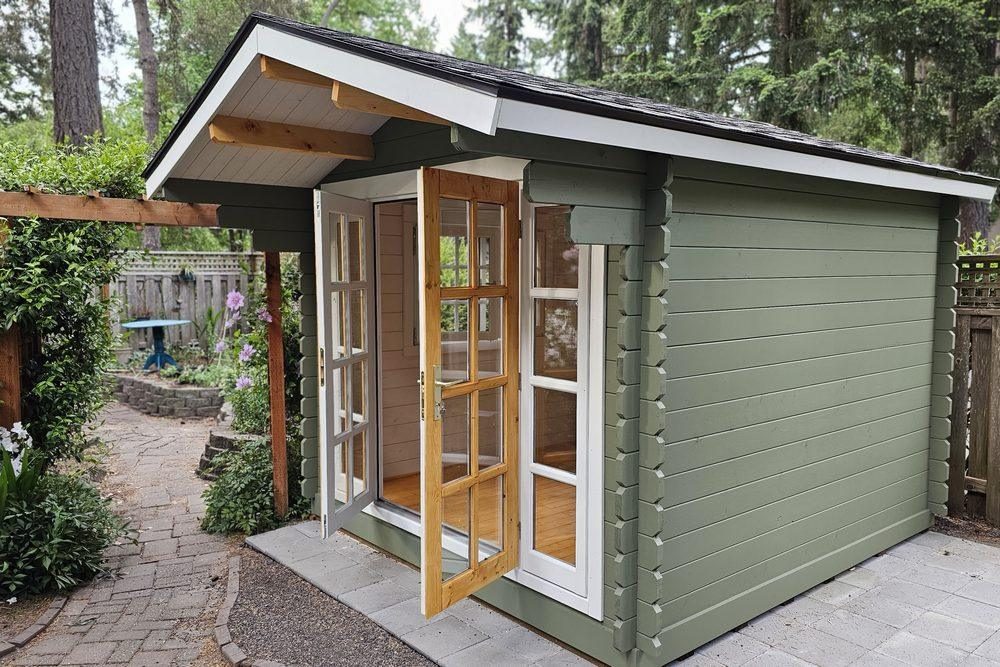 A sage-green Myrtle shed with French doors and an asphalt-shingle roof on a paver patio