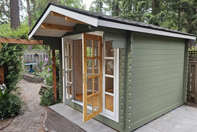 A sage-green painted Myrtle shed with French doors and a shingled roof, set on a paver patio in a customer's planted backyard