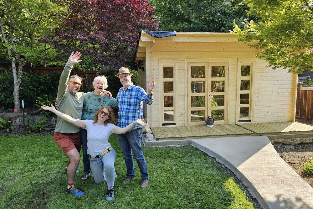 Four people cheering with their arms raised in front of a finished natural-wood Washington shed at the end of a weekend build