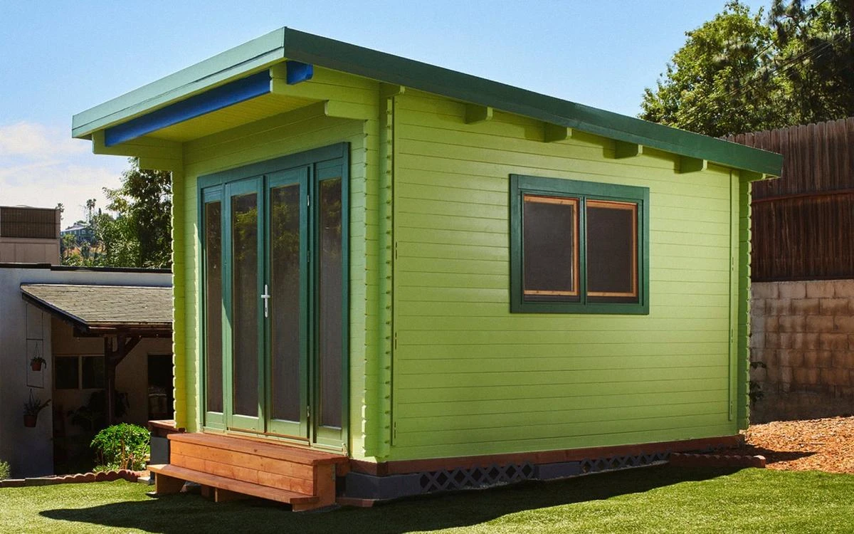 Paint preview tool: the California shed rendered in dark forest green with cream trim and an asphalt roof, beside the finish picker showing stain, neutral and color families
