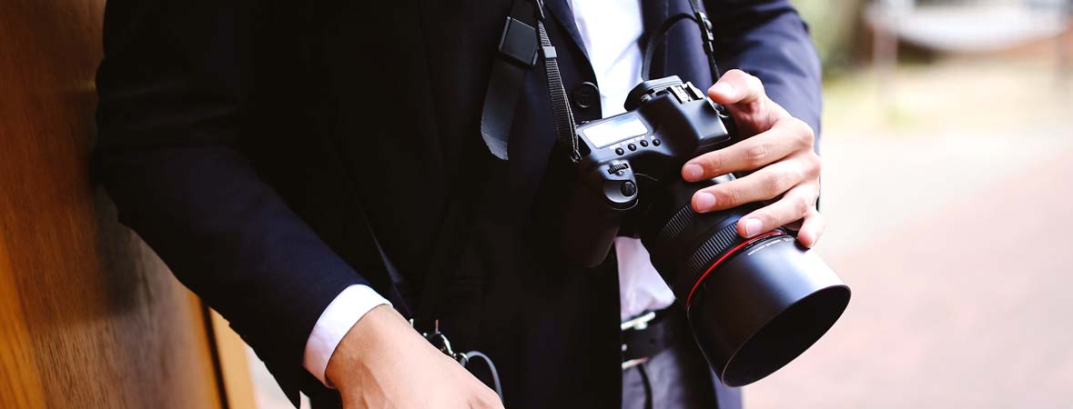 A photographer is dressed in a suit jacket and has a camera on a strap on his neck