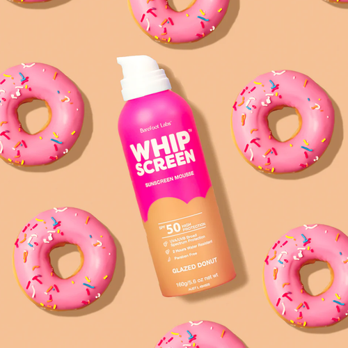A pink can of Barefoot Labs WHIPSCREEN Sunscreen Mousse with SPF 50 and "Glazed Donut" flavor, surrounded by pink frosted donuts with sprinkles.