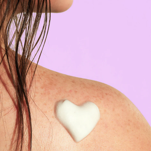 A white heart-shaped dollop of foam rests on a person's shoulder, which is covered in freckles. The background is a solid lavender color.