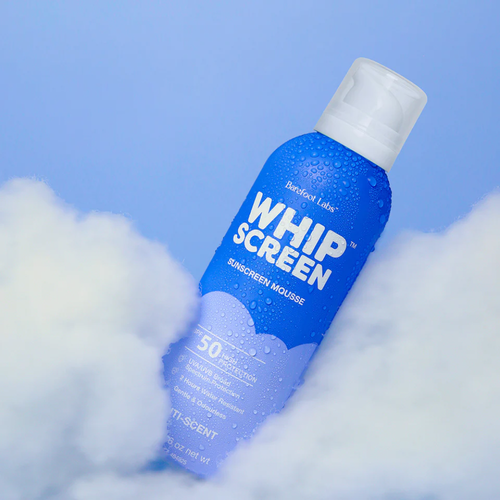 A blue bottle of Barefoot Labs Whip Screen Sunscreen Mousse with SPF 50 is surrounded by white clouds against a light blue sky. The bottle is covered in water droplets.