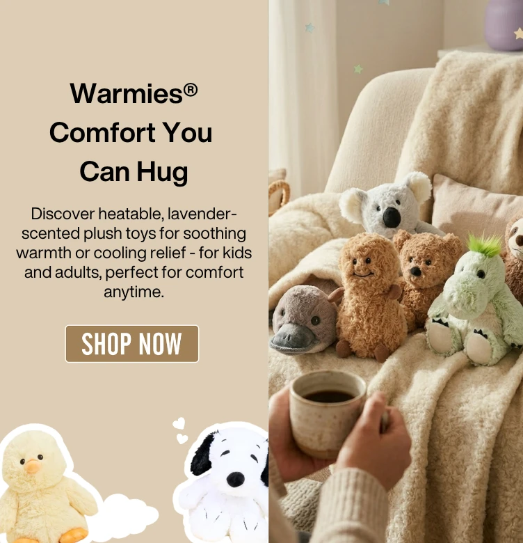 A digital promotional banner for Warmies plush toys. On the left, a tan background features the heading 'Warmies® – Comfort You Can Hug' followed by text describing heatable, lavender-scented plush toys for kids and adults. A 'SHOP NOW' button sits in the center, flanked by a yellow chick and a white dog plushie. On the right, a cozy lifestyle photo shows a group of Warmies—including a platypus, peanut, bear, koala, dragon, and sloth—nestled on a cream-colored armchair with a fuzzy blanket, while a person in the foreground holds a warm mug."