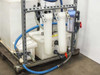 Lower portion of USFilter system showing three white filter housings, small secondary tanks, and blue hose connections on metal pallet frame