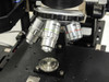 Olympus objective lens turret close-up — showing two Olympus MDPLAN objectives on turret with sample stage below and indenter tip holder