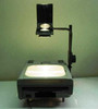Dukane 663 portable overhead projector shown fully deployed with projection head raised on arm and stage lamp lit — front view on table