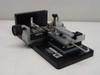 Applied Magnetics precision linear slide — three-quarter front-right view showing full assembly including right-angle end block, knurled adjustment knob, carriage with clamp, and micrometer barrel on base plate