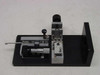 Applied Magnetics precision linear slide — three-quarter rear-left view showing Mitutoyo-style micrometer barrel extended, sliding carriage, spring post assembly, and black anodized aluminum base