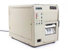 SATO M-8400 Industrial Barcode Label Printer front-right three-quarter view showing LCD display panel, indicator LEDs, and label exit slot with caution tag