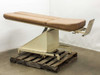 Tri W-G 645 chiropractic table in flat horizontal position, front three-quarter view showing tan upholstered surface, foot pedal on right side, and cream steel pedestal base