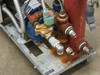Close-up of fluid manifold on AEC TDWF7M09S4 showing blue centrifugal pump, solenoid valve, brass fittings, water outlet port, and surface rust on piping