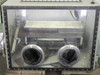 Vacuum Atmospheres glove box interior – front viewport view showing dual glove port openings with ripped black gloves, interior shelf, and stainless lining