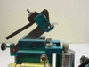Pneumatic actuator and measurement stage – angled view showing teal anodized aluminum body, pivot arm, and pneumatic cylinder