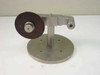 Custom Scientific stainless steel component holder stand — front-right view showing round bolt-down base, vertical post, adjustable pivot arm, and dark abrasive or rubber disc mounted on center hub