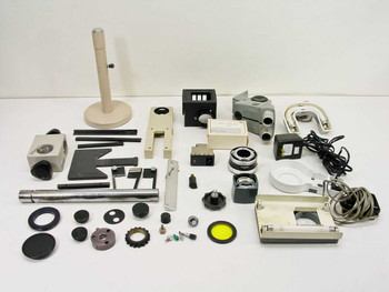 Full overhead view of approximately 36 mixed microscope parts spread on white surface including stand, heads, filter cubes, ring lights, knobs, and adapters