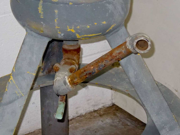 Bottom base area of APHD 10STDWPB tank showing rusted drain pipe fitting with open-end pipe stub and floor stand legs