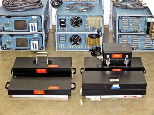 Four HNVS Test Fixture Assemblies and two stacked blue chassis units — top-down view of fixture trays with pin grid arrays and orange warning labels; system rear panels with BNC ports visible in background