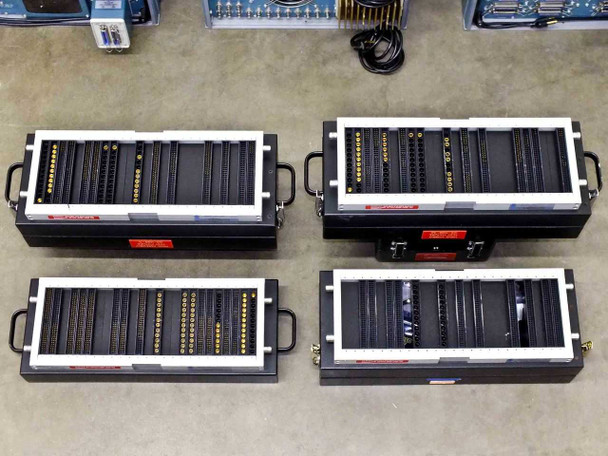 Four HNVS Test Fixture Assemblies laid out — top view showing all four trays with dense pin/socket grid arrays, orange hazard labels, carry handles, and connector strips