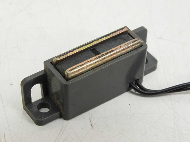 Close-up of magnetic proximity reed switch body showing gray plastic housing, exposed reed contact elements, and mounting flanges with screw holes