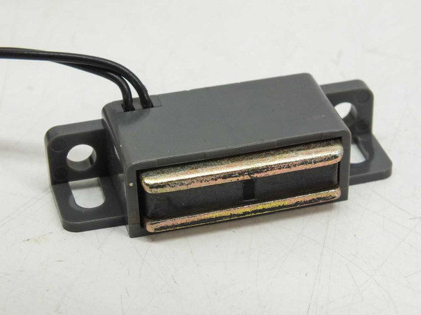 Side view of magnetic proximity reed switch showing rectangular gray housing, metallic reed contact strips, and short black lead wire exiting rear