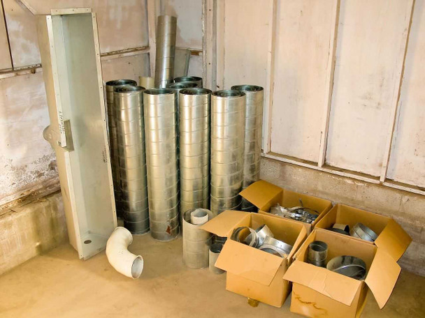 HVAC galvanized sheet metal round duct sections stacked by diameter with sheet metal basin at left and four cardboard boxes of fittings at right