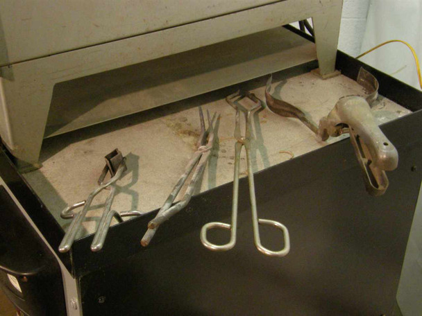 Furnace accessory tools laid out on cabinet shelf — includes tongs, crucible tongs, and a heat-resistant glove visible among the tools