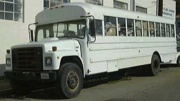 1987 International S1800 32-foot white passenger bus parked outside warehouse, driver-side front three-quarter view showing full body length
