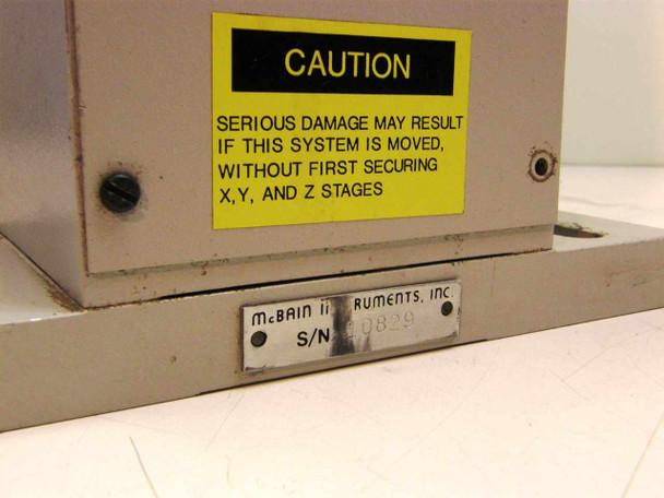 Close-up of McBain Instruments nameplate with serial number and yellow caution sticker warning to secure X, Y, and Z stages before moving