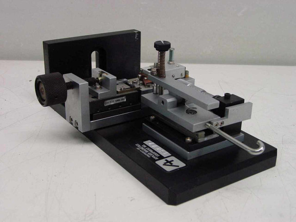 Applied Magnetics precision linear slide — three-quarter front-right view showing full assembly including right-angle end block, knurled adjustment knob, carriage with clamp, and micrometer barrel on base plate