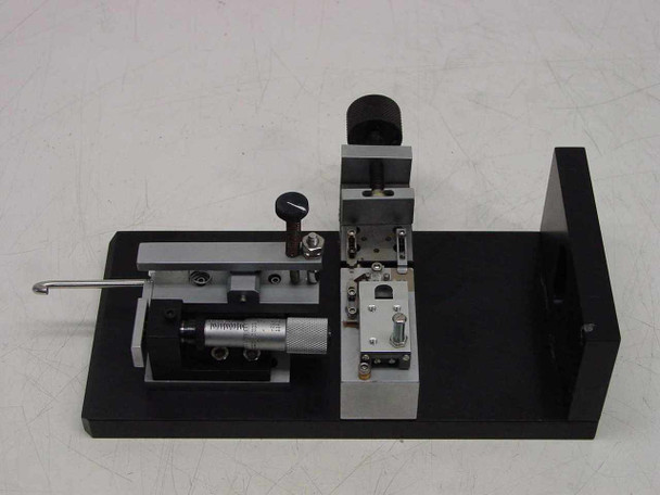 Applied Magnetics precision linear slide — three-quarter rear-left view showing Mitutoyo-style micrometer barrel extended, sliding carriage, spring post assembly, and black anodized aluminum base