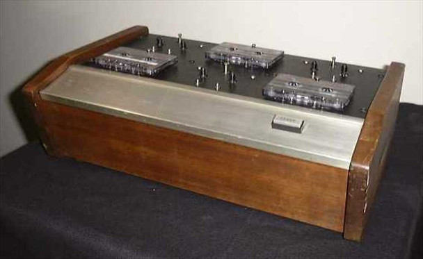 Pentagon CW6 6-tape high-speed audio cassette rewinder — three-quarter front view showing wooden housing, brushed metal top deck with six cassette bays, three cassettes loaded, and front control button