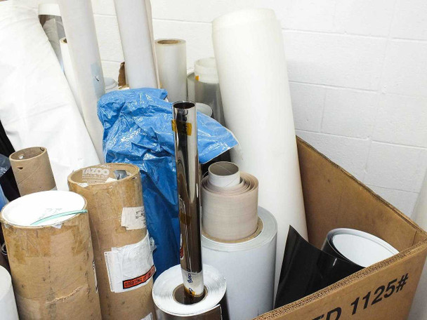 Side-angle view of film rolls including chrome tube, white film rolls, brown cardboard core rolls with label partially reading TAZOO, and a black film roll at far right
