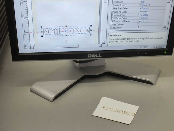Dell monitor displaying Synrad WinMark Pro software with RECYCLEDGOODS.COM text layout, with physical laser-marked sample card in foreground showing completed marking