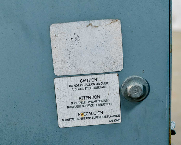HPS NMF025LE transformer – close-up of caution label reading 'Do Not Install On or Over a Combustible Surface' in English, French, and Spanish, with mounting bolt visible