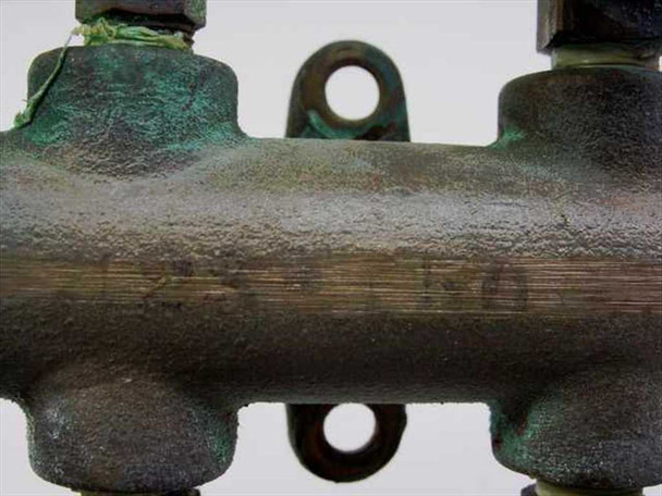 Close-up of brass manifold body showing surface patina, verdigris oxidation, and stamped markings on the barrel