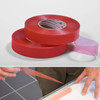 The image shows a pair of hands applying ORABOND® 1397PP Double-sided Adhesive Tape to a large vinyl banner. The tape, with its red PP film release liner, is being carefully aligned along the edge of the banner. The hands are pressing down firmly to ensure a strong bond. The banner itself is laid out on a flat surface, with vibrant colors and text indicating its use for signage. The background shows a clean workspace with tools and materials neatly arranged, highlighting a professional environment. The focus is on the precision and quality of the application process, emphasizing the tape’s role in securing the banner effectively.