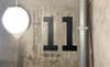 A concrete wall inside a modern industrial-style building displays large black vinyl numbers “11” along with smaller text reading “FLOOR” above and “FLATS 94–101” below. The signage provides clear directional information, illuminated by a soft round light fixture to the left and accented by exposed metal piping on the right.