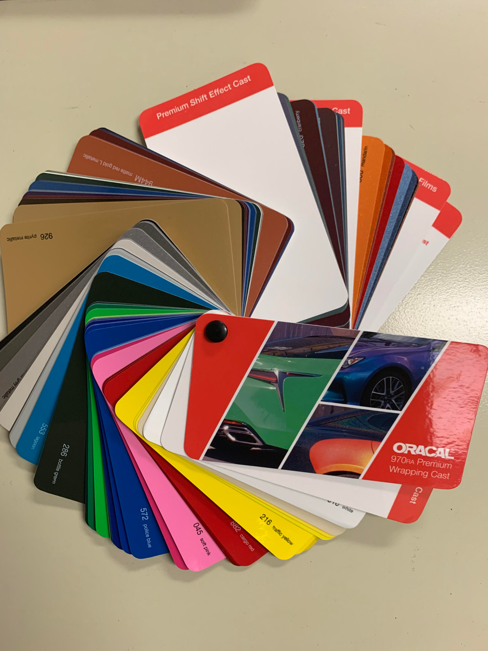 The image showcases the ORAFOL ORACAL 970RA Sample Swatch Deck, a compact and professional tool for vehicle customization. The swatch book is open, displaying a fan of 80 vibrant color samples, including glossy, matte, and metallic finishes. Each sample is precisely cut and labeled, allowing for easy comparison and selection. The sleek, organized design of the swatch deck highlights its high-quality construction and practicality for both professional and DIY use. The background is neutral, ensuring the vivid colors of the swatches stand out prominently.