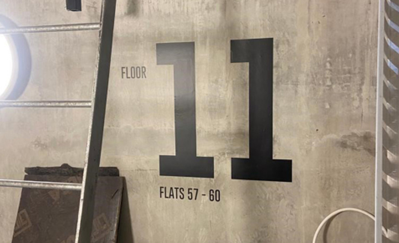 A concrete wall inside a modern industrial-style building displays large black vinyl numbers “11” along with smaller text reading “FLOOR” above and “FLATS 94–101” below. The signage provides clear directional information, illuminated by a soft round light fixture to the left and accented by exposed metal piping on the right.