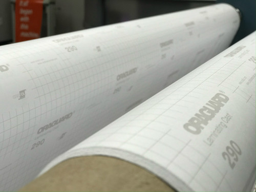 The image showcases a modern, professional laminator in the process of applying the Orafol ORAGUARD® 290F Optically Clear LAMINATE. The laminate roll, characterized by its 2-mil thickness and gloss finish, is mounted above the machine. Below, a large-format digital print passes through the laminator, where the clear, permanent adhesive of the ORAGUARD® 290F seamlessly adheres to the print's surface, enhancing its durability and UV protection. The scene is set in a well-lit workspace, emphasizing the clarity and smooth application of the laminate, free from air bubbles and wrinkles.