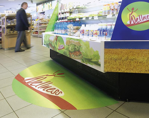 This image captures a vibrant, full-color printed advertisement strategically placed on the floor of a grocery store deli. Positioned right in front of the counter, the advertisement showcases a promotional offer, engaging customers as they wait to be served. The graphic is printed using Mactac PERMACOLOR FloorGrip, highlighting its clarity and durability in a high-traffic area.