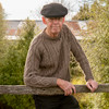 Man leaning on fence wearing Lesley Unisex Aran Wool sweater in the colour Brown.